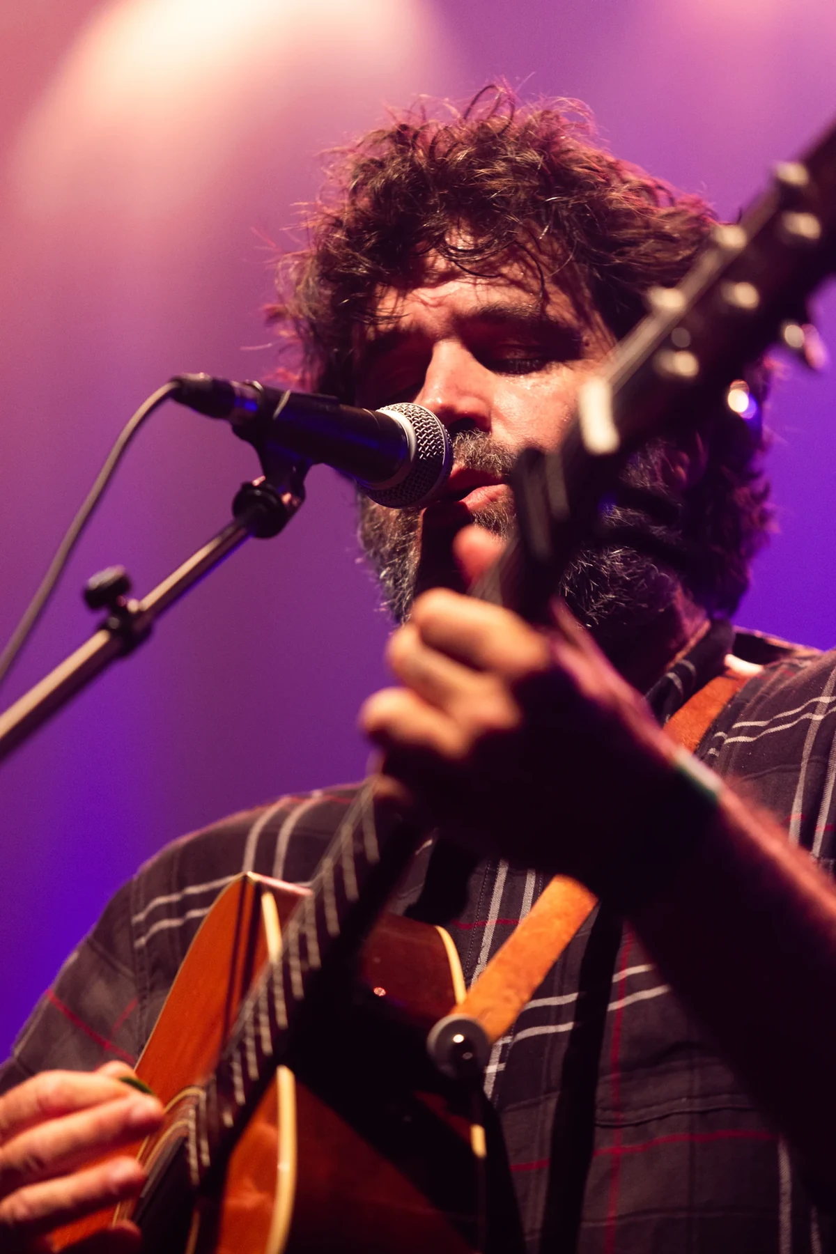 Portrait rapproché du chanteur de La Maison Tellier au micro, tenant sa guitare sous une lumière rose.