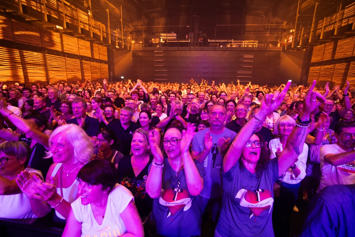 Le public du concert de La Maison Tellier applaudit face à la scène, sous une lumière violette et dorée.