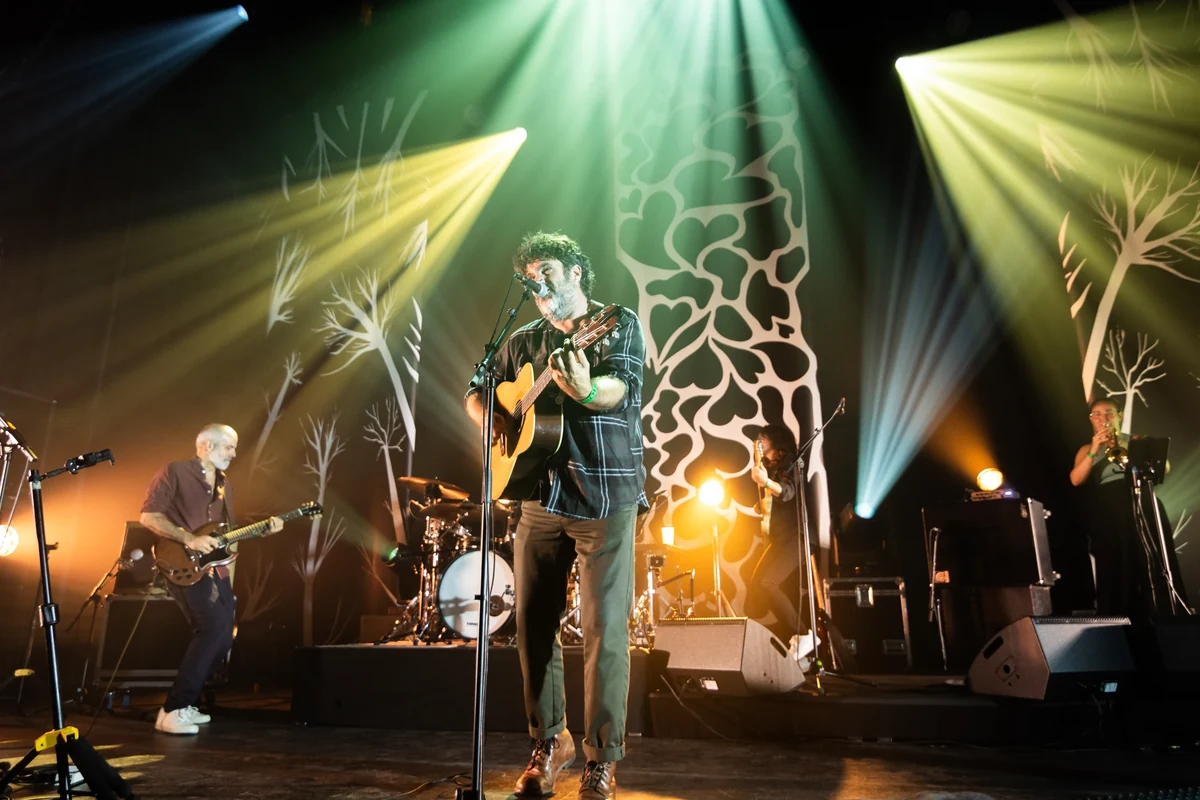 La Maison Tellier en concert, avec le chanteur à la guitare acoustique au centre et les musiciens sous des faisceaux colorés.