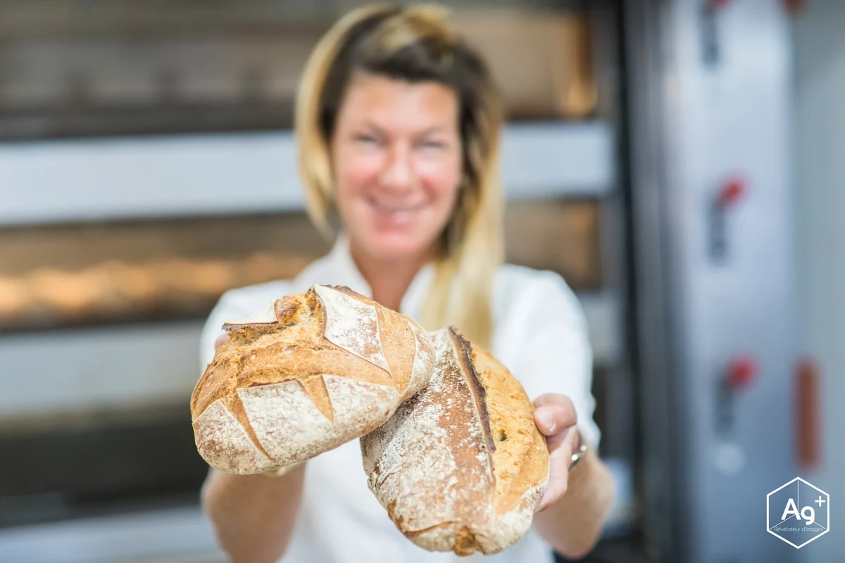 Une boulangère présente un pain rond vers l’objectif, devant le four de la boulangerie.