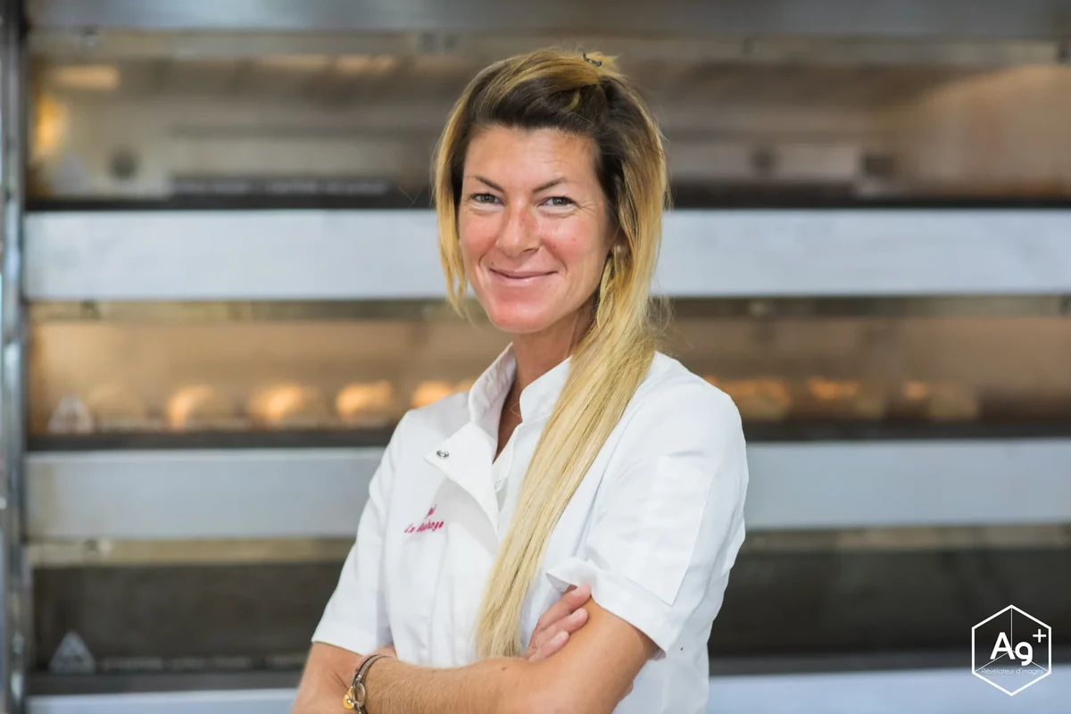 Une boulangère en tenue blanche, les bras croisés devant un four.